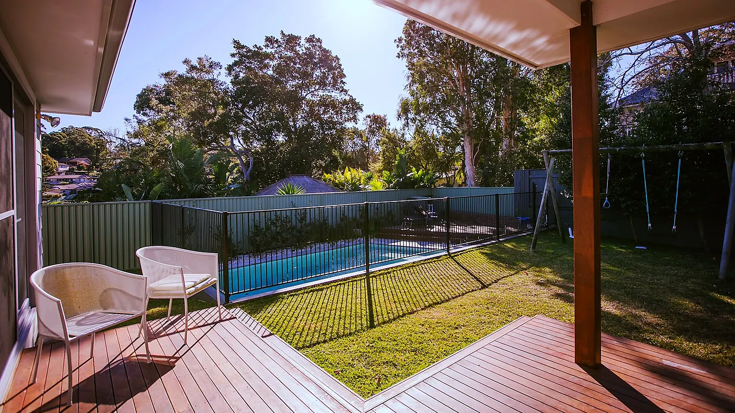 A covered timber deck with outdoor chairs, looking across lawn to a fenced pool and bushland beyond.