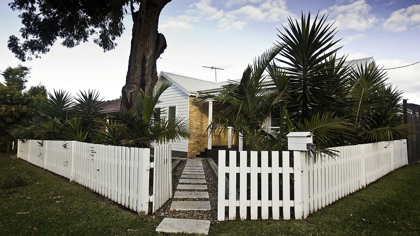 A white picket fence and gate opening to a paved path leading to a weatherboard cottage behind established trees.