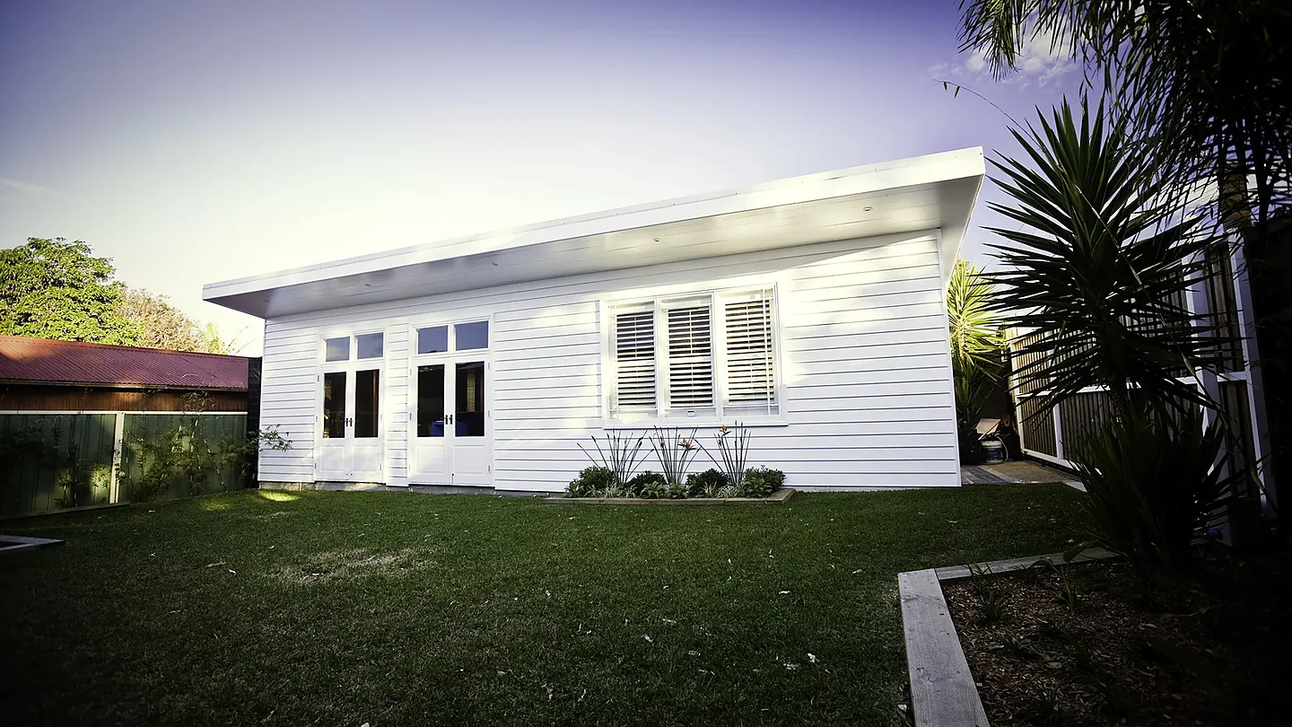 The rear elevation of a white weatherboard cottage with a small table on the lawn and planting to the boundary.