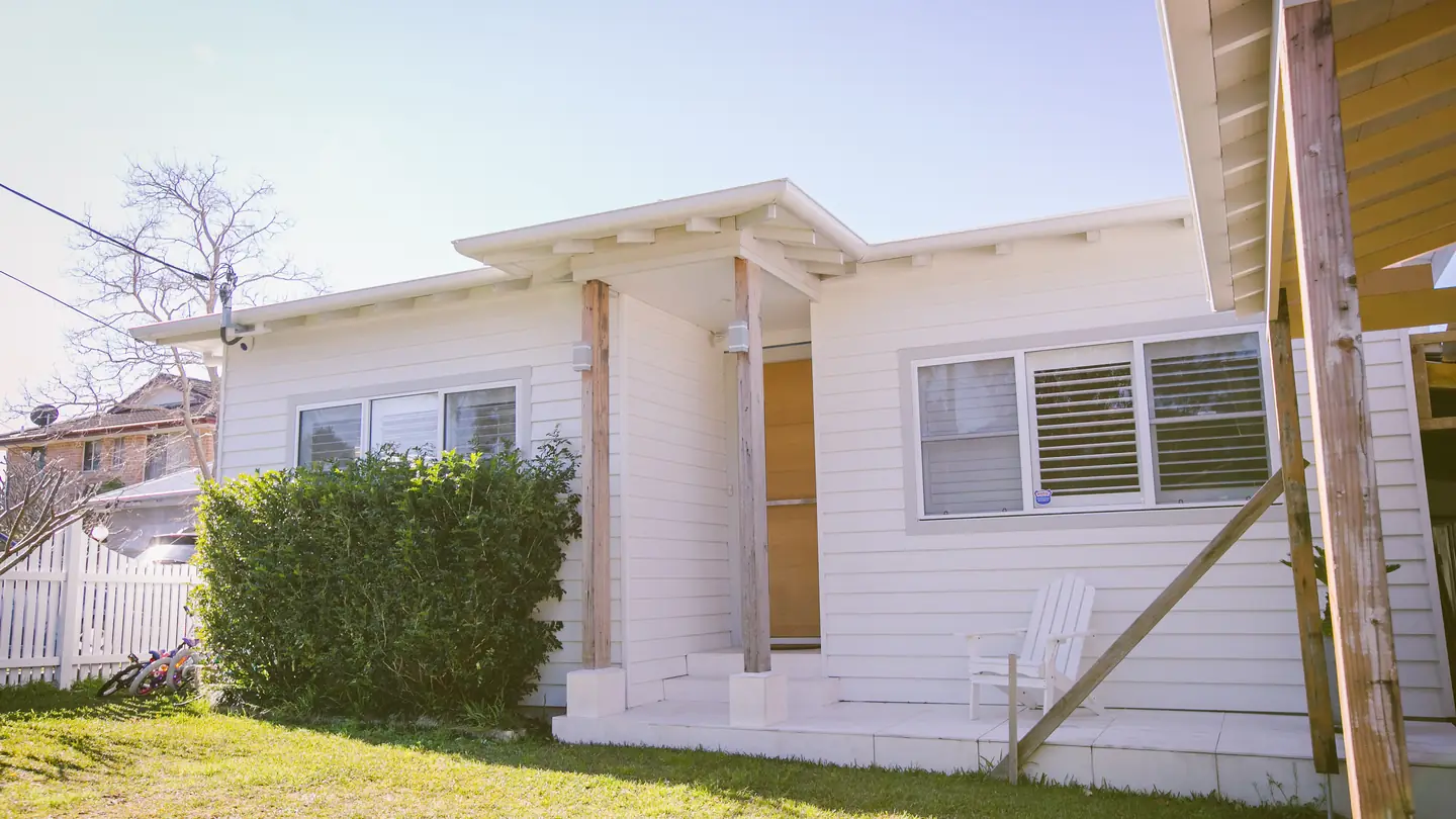 The front of a white weatherboard cottage with a timber-posted porch and clipped hedge.