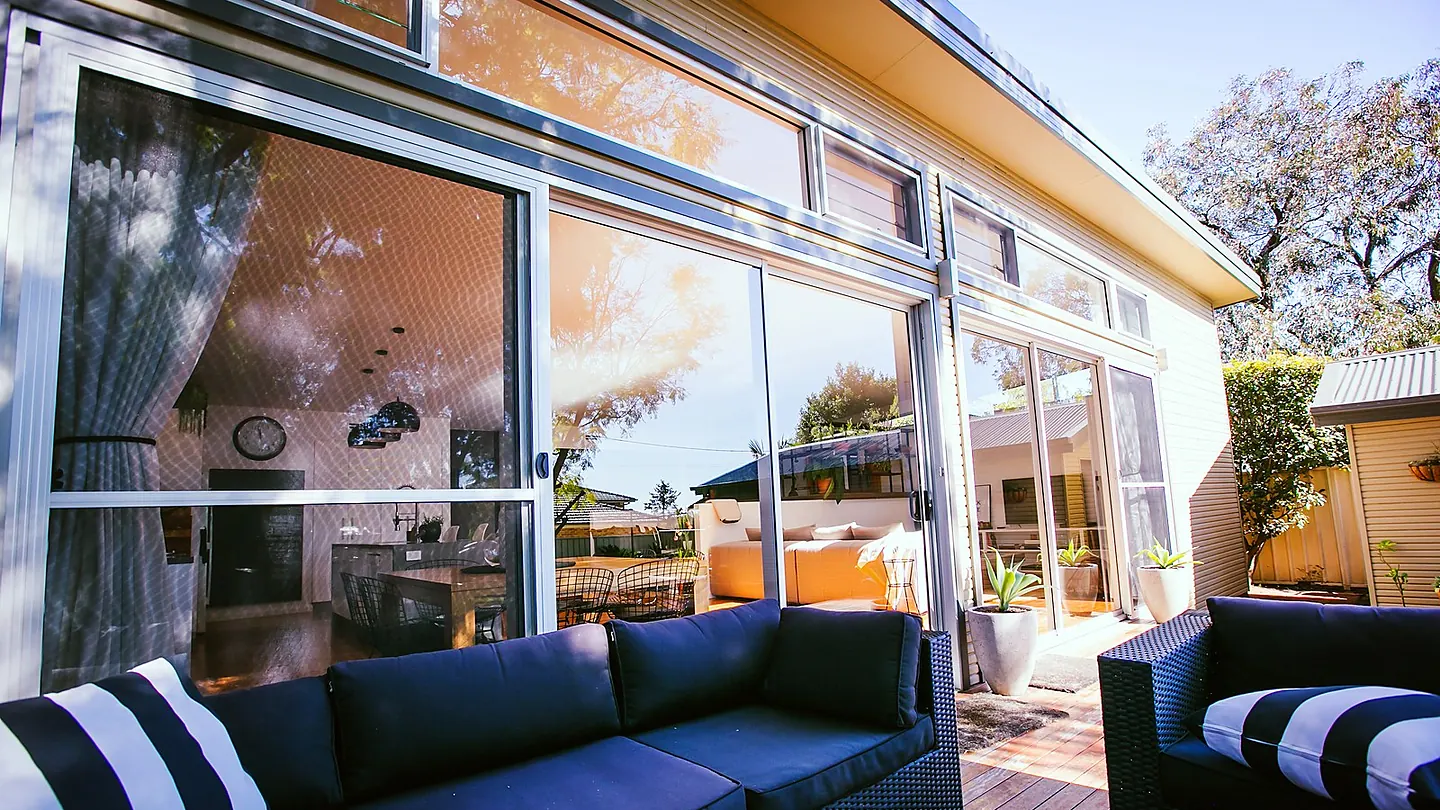 An outdoor lounge setting on a timber deck, with full height glazing reflecting the garden behind.