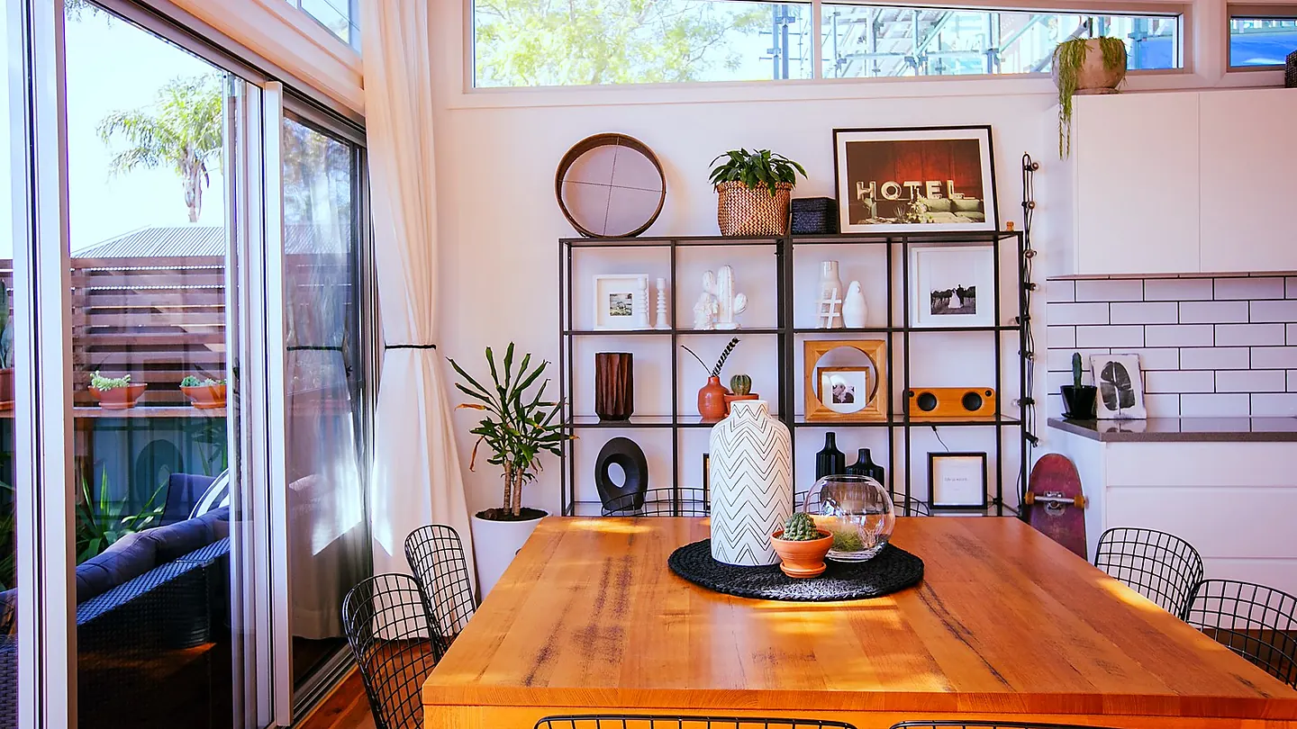 A timber dining table beside open shelving, with sliding doors to a deck and a brick splashback behind.