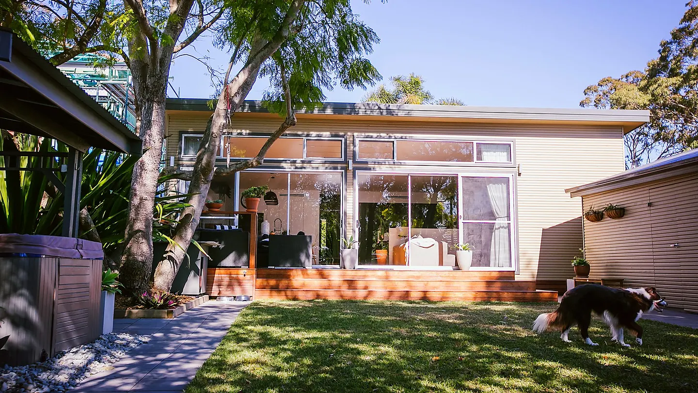 A rear garden with lawn and established trees, looking back to a low clad house with a pergola along its length.