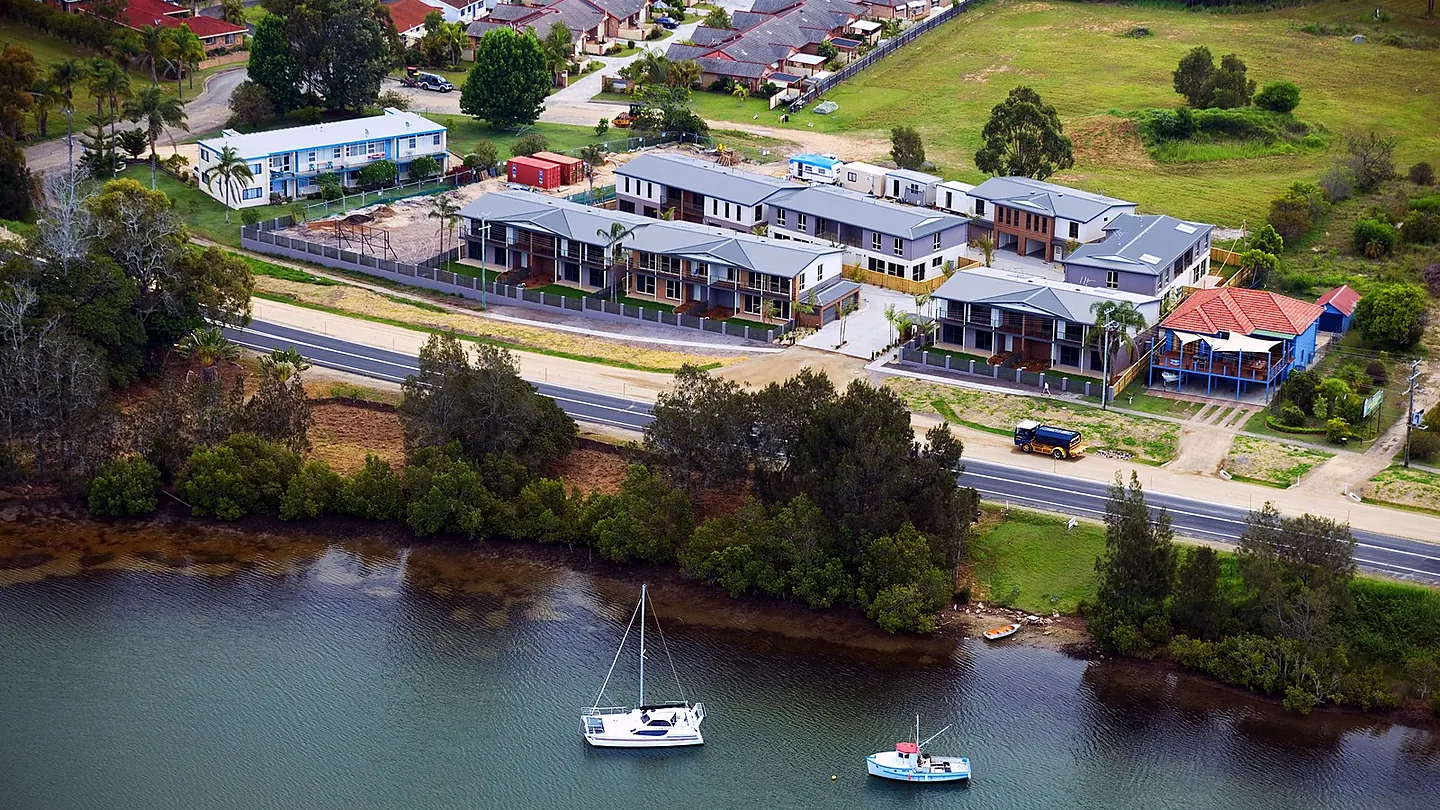 An aerial view of a row of single and two storey dwellings along a waterfront road, with moored yachts in the channel.