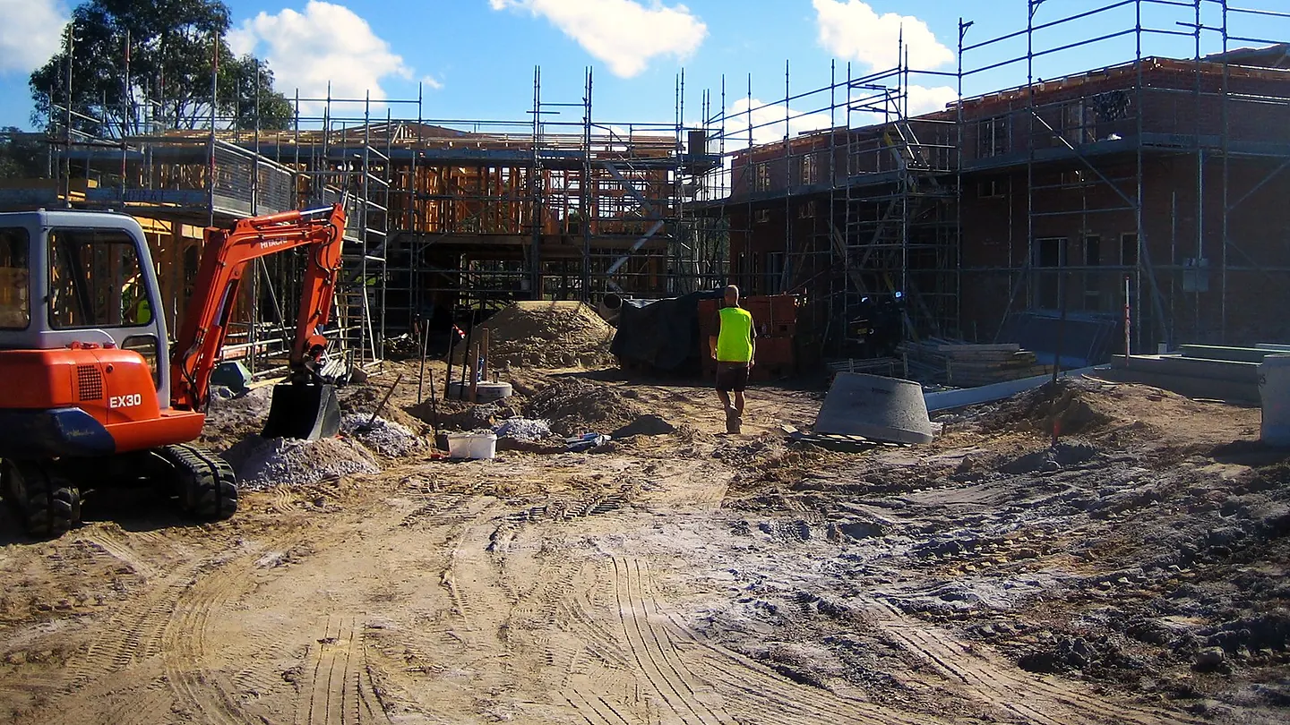 An excavator working on a site at slab stage, with scaffold to the structure behind and spoil stockpiled in the foreground.