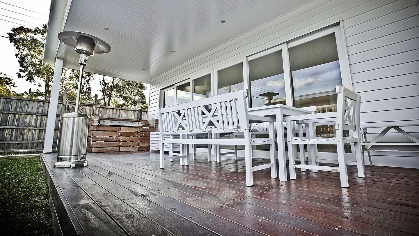A covered timber verandah with a long table and white chairs, looking out through glazed doors.
