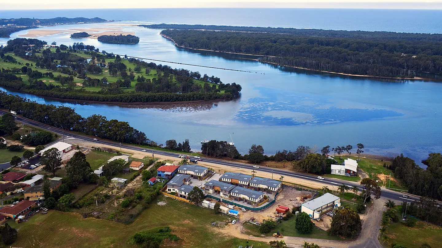 An aerial view of a coastal town on a river, with an island midstream and a development site in the foreground.
